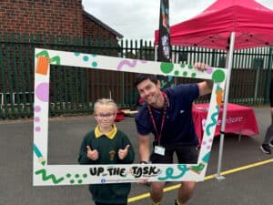 A child and an adult are posing for a photo while holding a large, colorful rectangular frame with the text "Up for the Task". They are standing outside near a red tent and a brick building, giving thumbs-up gestures and smiling at the camera.